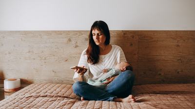 descriMother on bed with newborn on her mobile doing life admin and personal tasksption