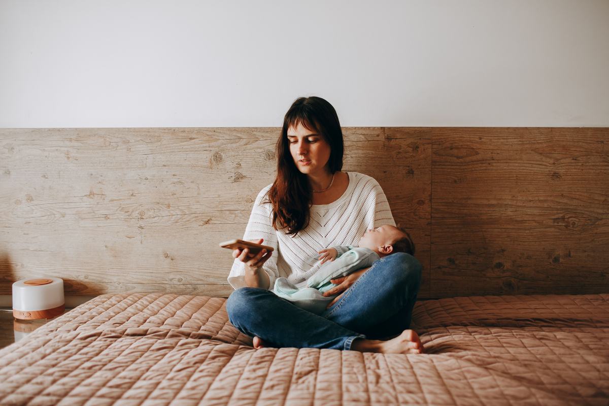 descriMother on bed with newborn on her mobile doing life admin and personal tasksption